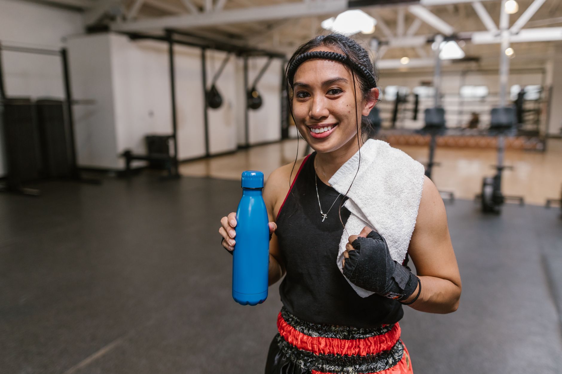 Asian woman in gym holding a water bottle and towel, smiling confidently.