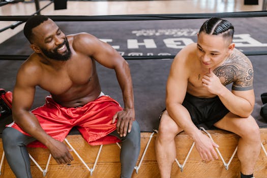 Two shirtless athletes sitting on a gym bench, smiling and relaxing after training.