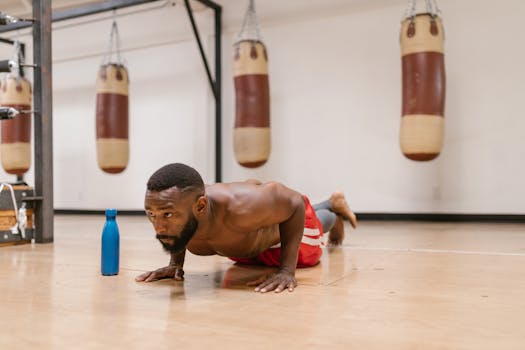 A focused shirtless man performing push-ups on a gym floor beside a blue water bottle.
