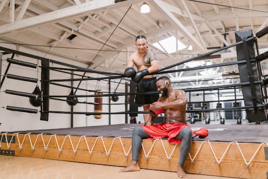 Two men in a boxing ring, smiling after a workout. One is sitting, the other standing.