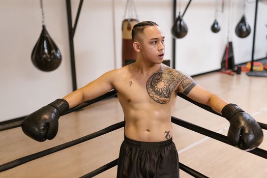 Shirtless male boxer with tattoos and gloves resting against ropes in a gym.