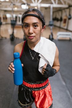 Young female athlete in gym gear resting after workout, holding a blue water bottle and towel.