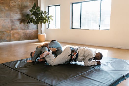 Two men in martial arts uniforms practicing Brazilian Jiu-Jitsu on gym mats indoors.