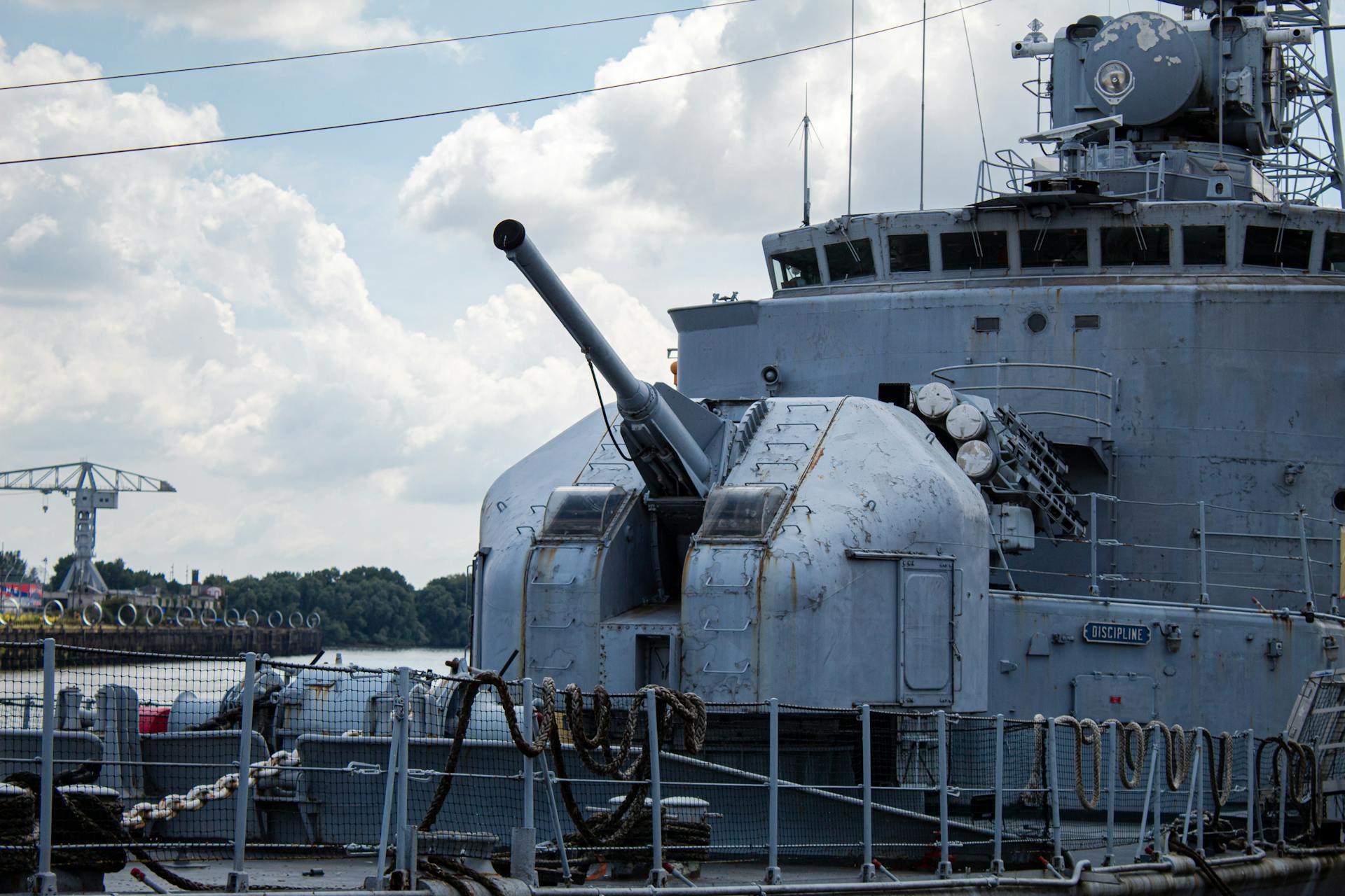 Close-up of a naval warship cannon on deck at a harbor, showcasing its heavy machinery.