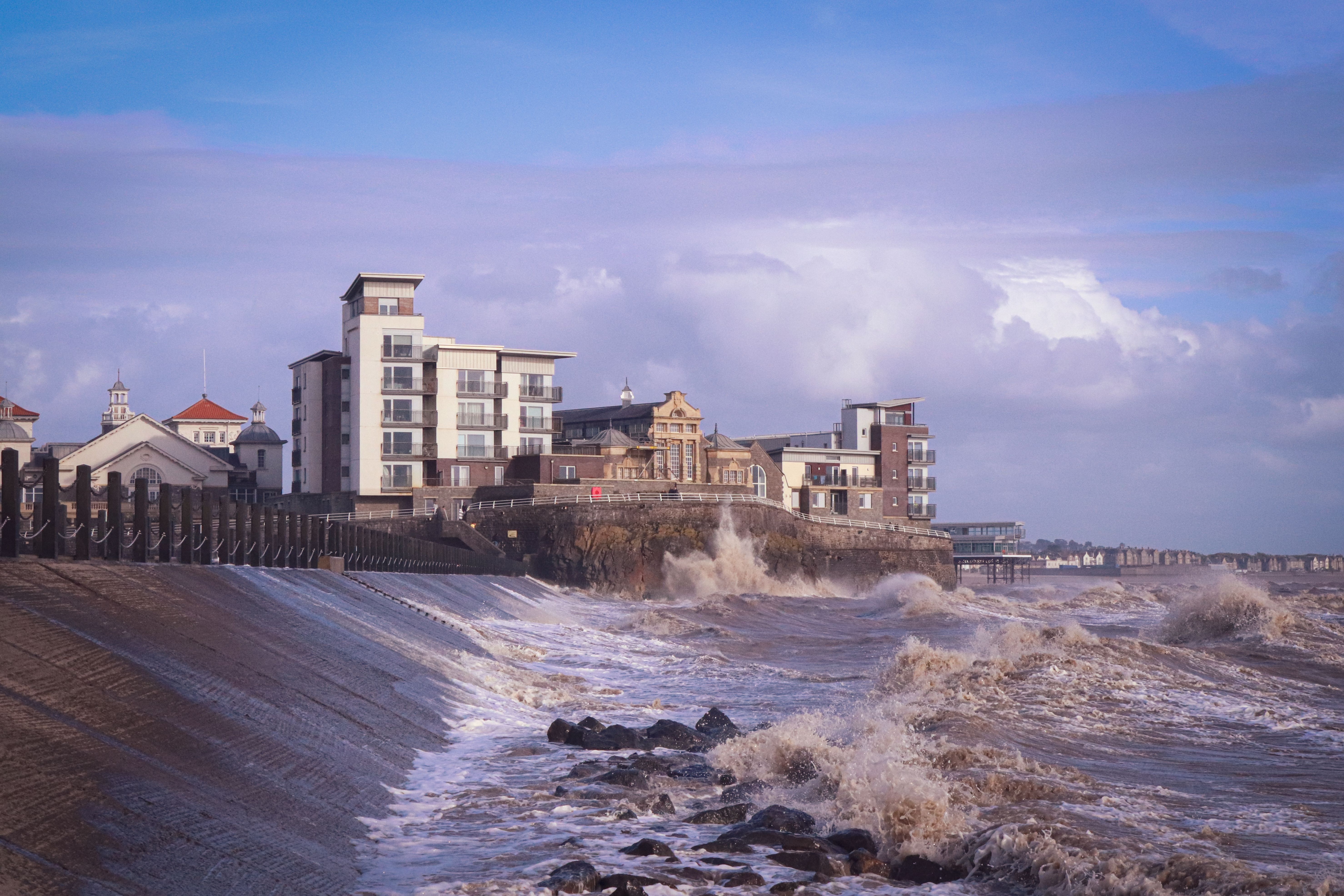 Buildings overlooking the sea with waves crashing on rocks and a clear sky.
