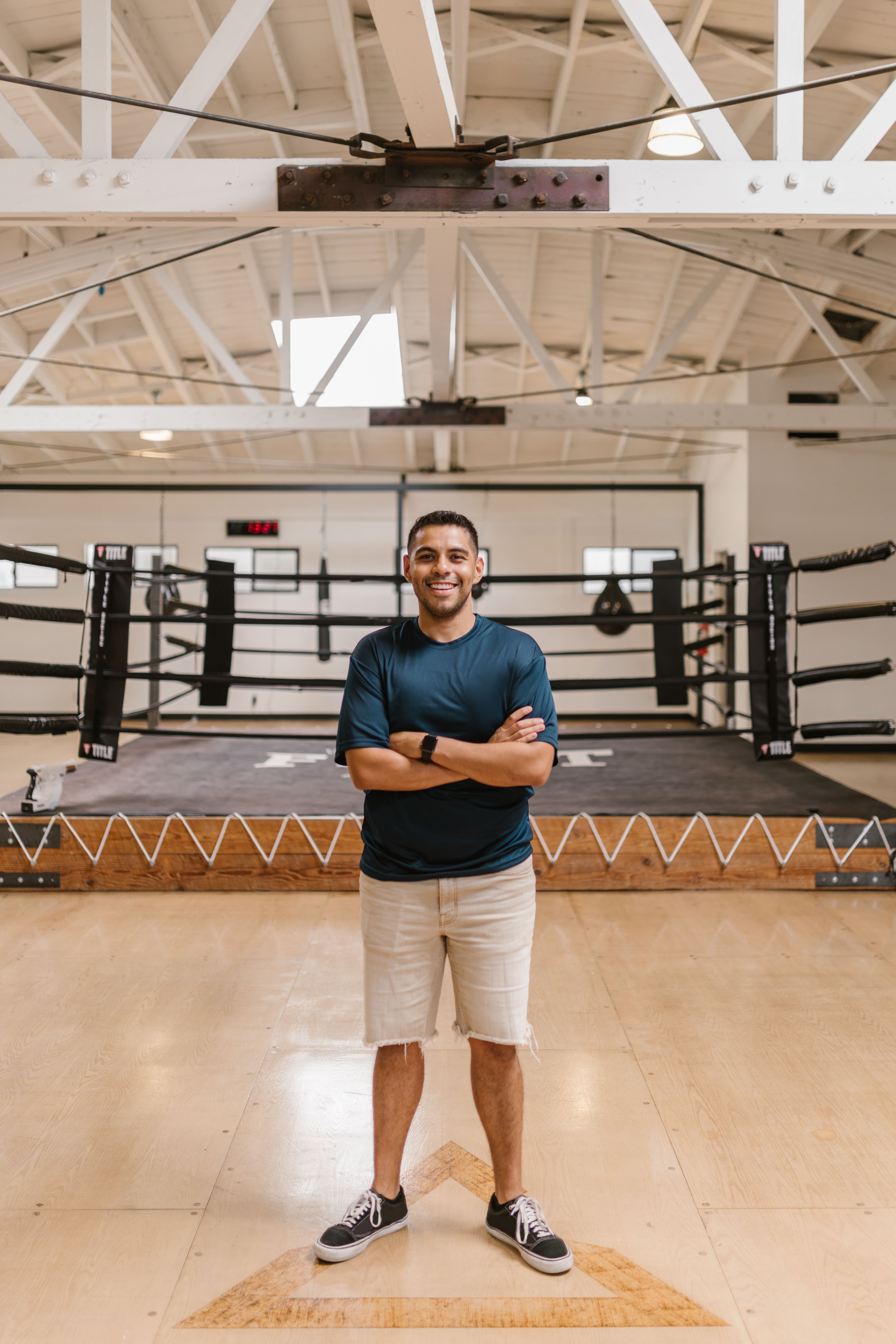 Man standing confidently in a boxing gym, wearing casual clothes with a boxing ring in the background.
