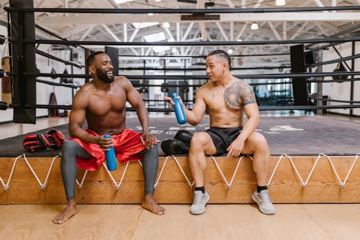 Two shirtless men sitting in a boxing gym, holding water bottles and smiling.