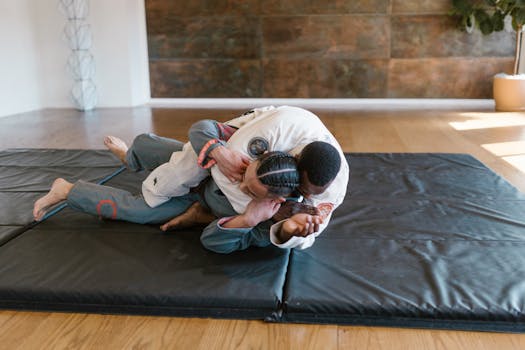 Two martial artists practicing grappling techniques on gym mats indoors.