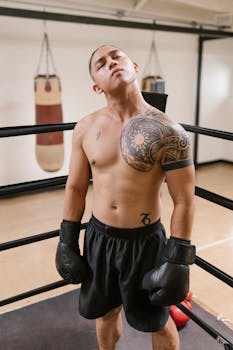 A muscular topless man with tattoos poses confidently in a boxing ring wearing gloves.