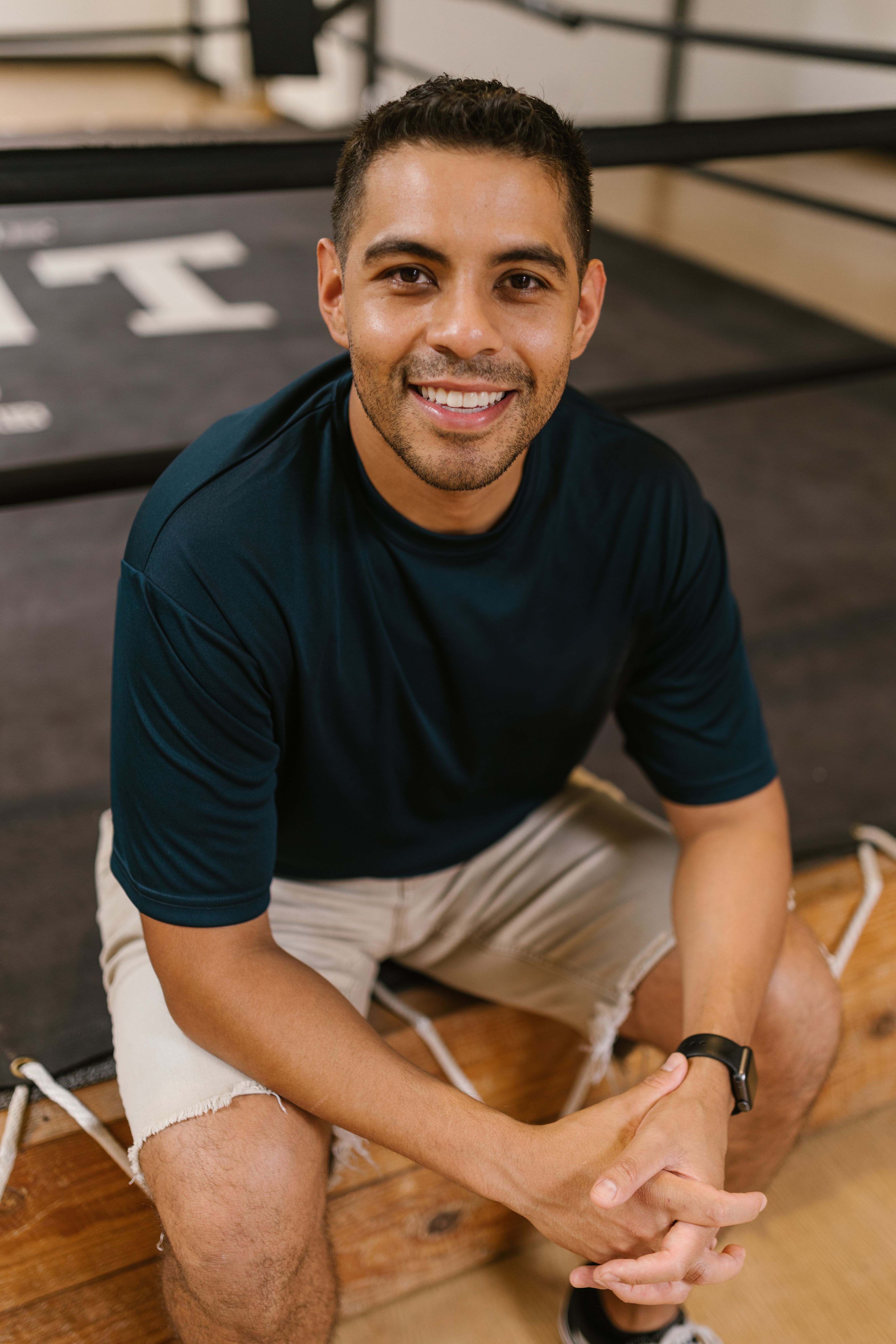 Cheerful young man with short hair sits by a boxing ring indoors, wearing a t-shirt and shorts.