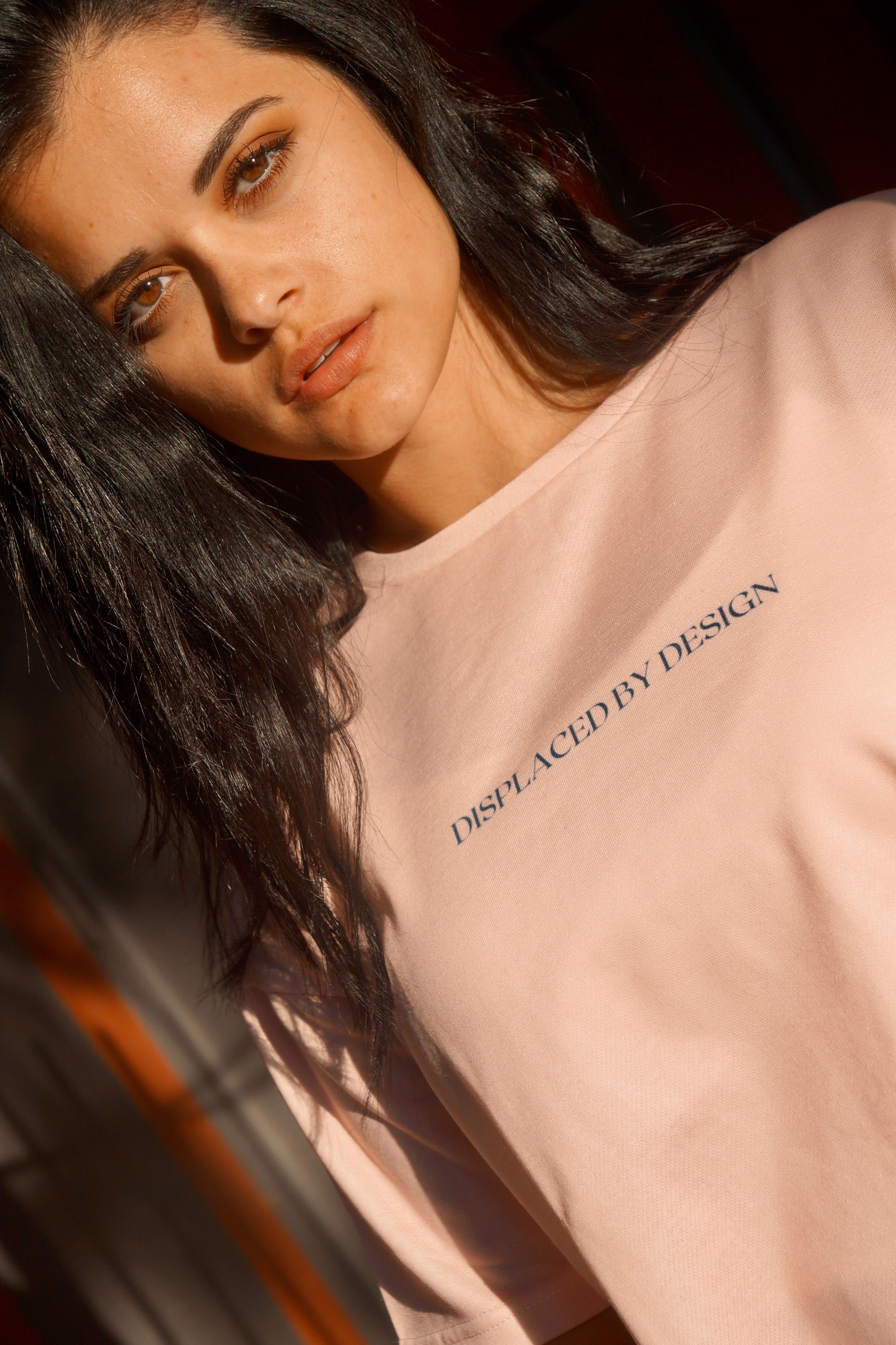 A woman wearing a pink shirt stands in the sunlight, displaying the motto 'Displaced by Design'.