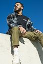 Man in Black and White Plaid Dress Shirt and Brown Pants Sitting on White Concrete Wall