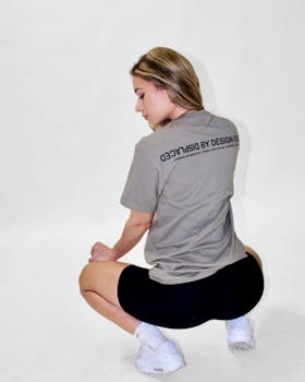 A woman squats in a casual outfit, showcasing a graphic tee in a minimalist studio setting.