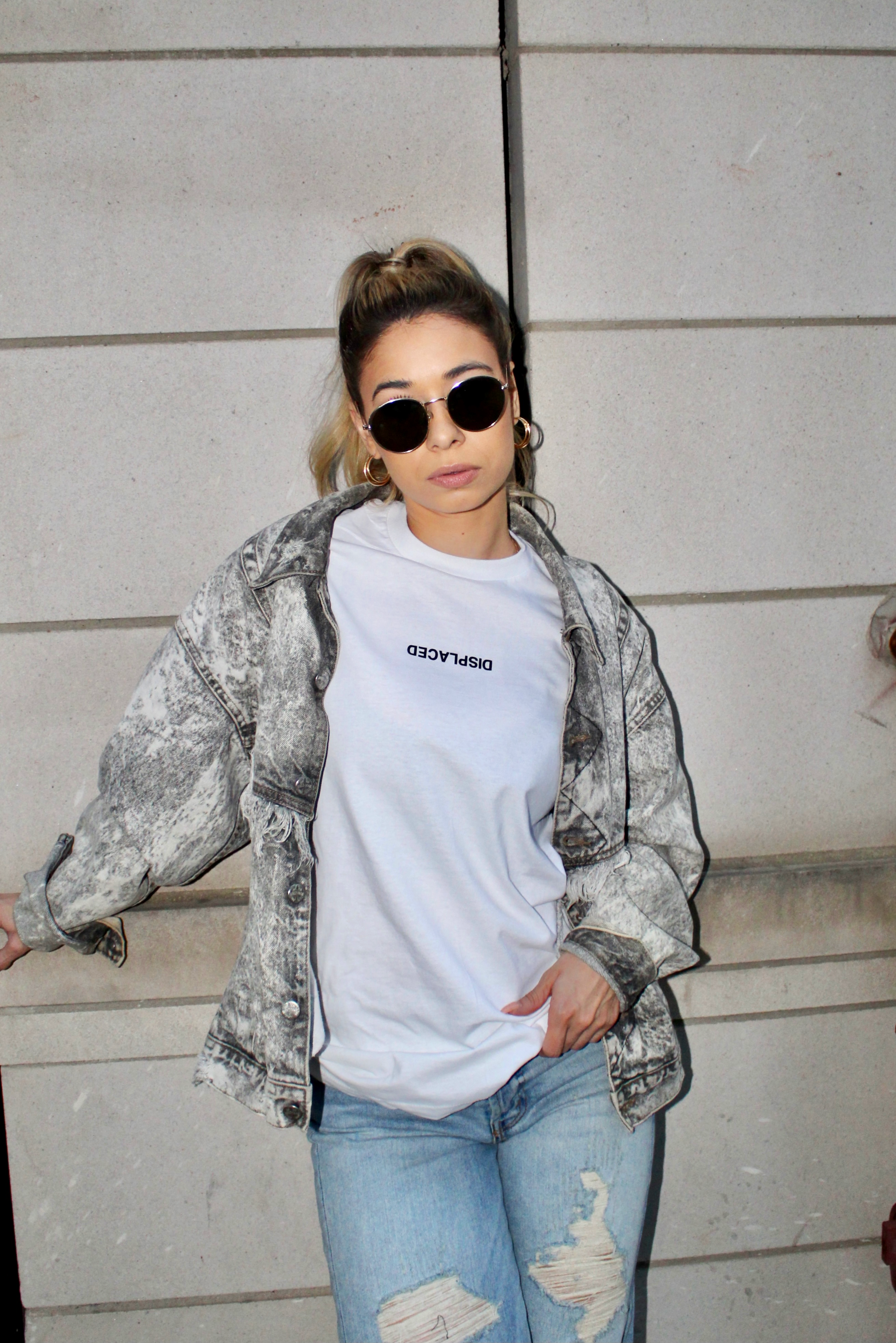 Stylish woman in urban streetwear with sunglasses and casual demeanor against a concrete wall.