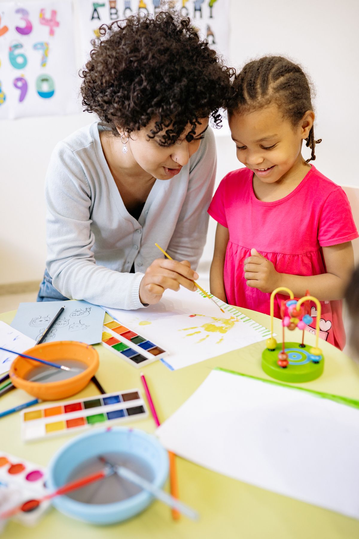 Teacher helping a young girl paint with watercolours at a classroom table