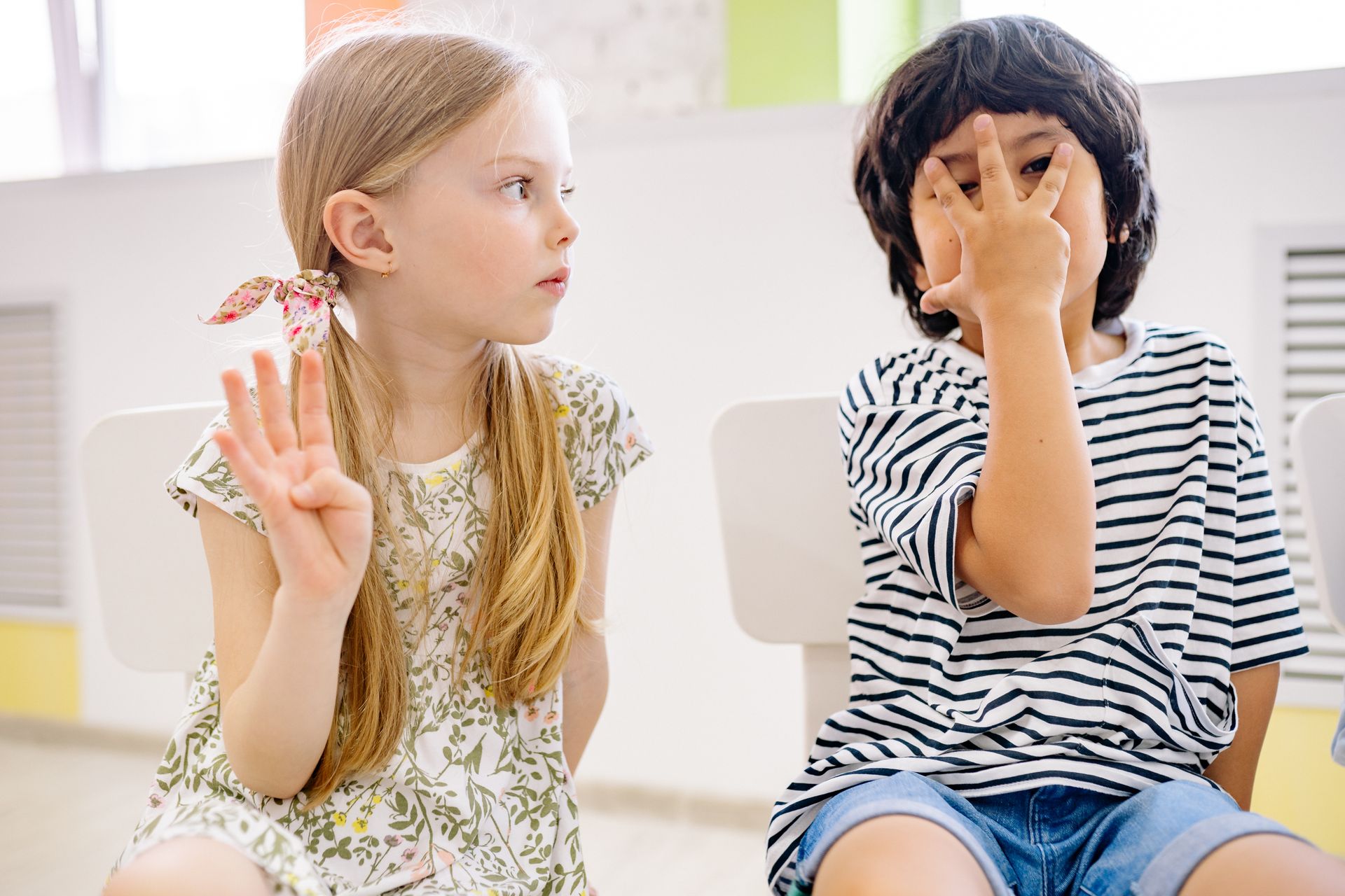 Two children sitting indoors engaging in a playful learning activity, showing numbers with fingers.