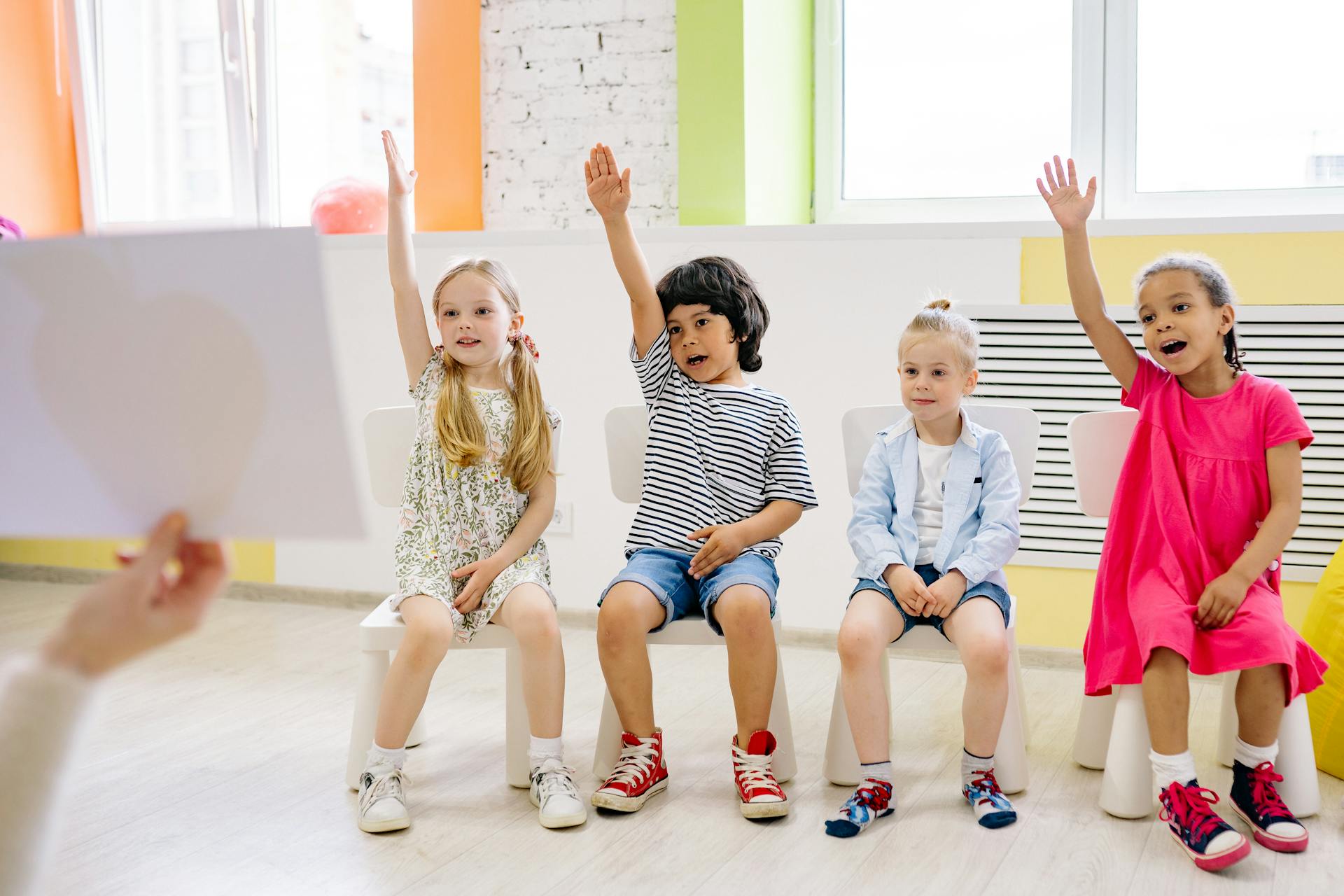 Four children eagerly raising their hands in a vibrant and colorful classroom setting.