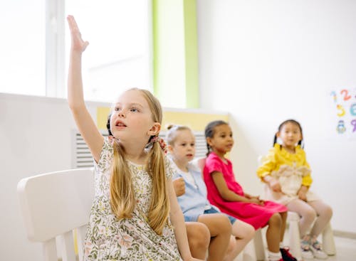 Girl in White and Black Floral Dress Sitting on Chair Raising Her Hand