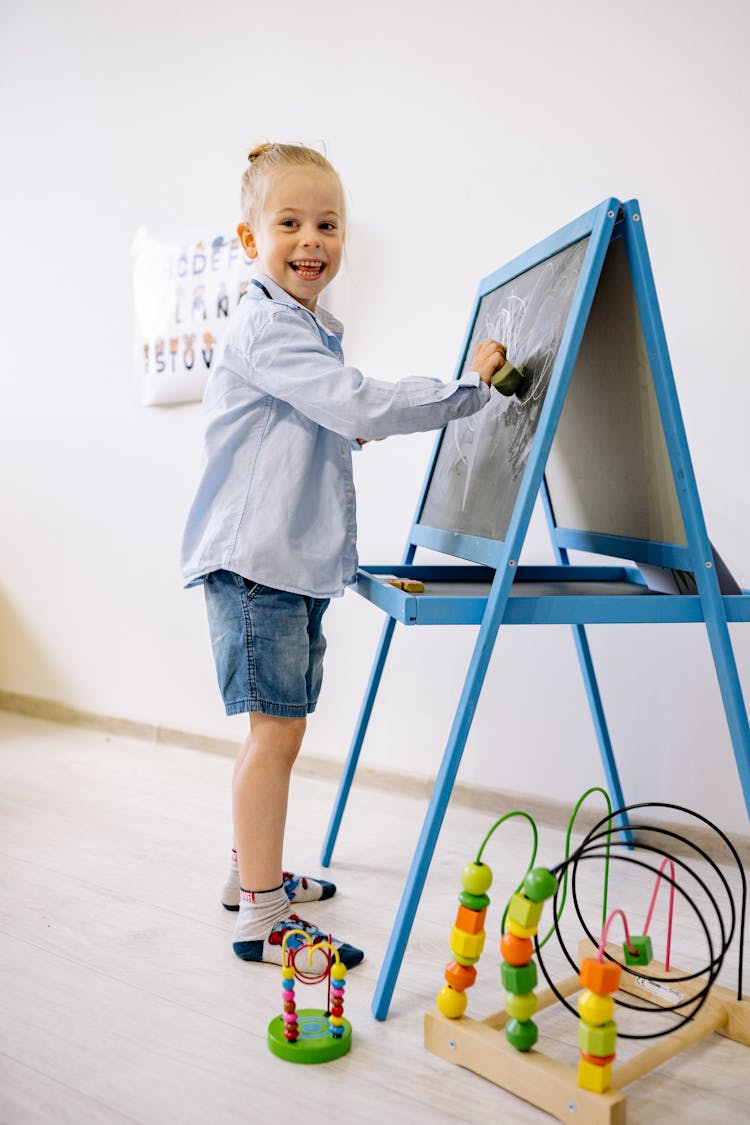 Child In Blue Long Sleeve Shirt And Blue Denim Shorts Learning To Draw On Blackboard