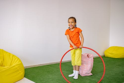Little Girl In Orange Shirt And Yellow Pants Playing With Hula Hoop