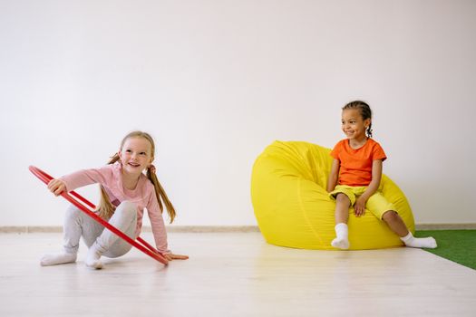 Two children enjoying playtime indoors with a hula hoop and bean bag, full of joy and fun.