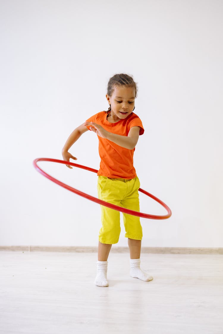 Little Girl In Orange Shirt And Yellow Pants Playing With Hula Hoop