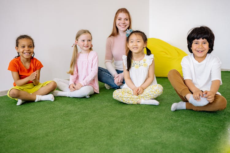 Happy Children And Teacher Sitting On Green Grass Carpet