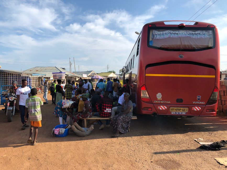 A busy scene at a Ghanaian bus station with locals gathered around a parked bus on a sunny day.