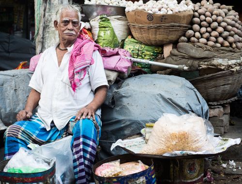 Free Senior vendor sitting with baskets of garlic and potatoes at a vibrant street market. Stock Photo