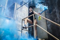 一名女子手持蓝色烟雾弹，站在工业楼梯上，营造出前卫的时尚场景