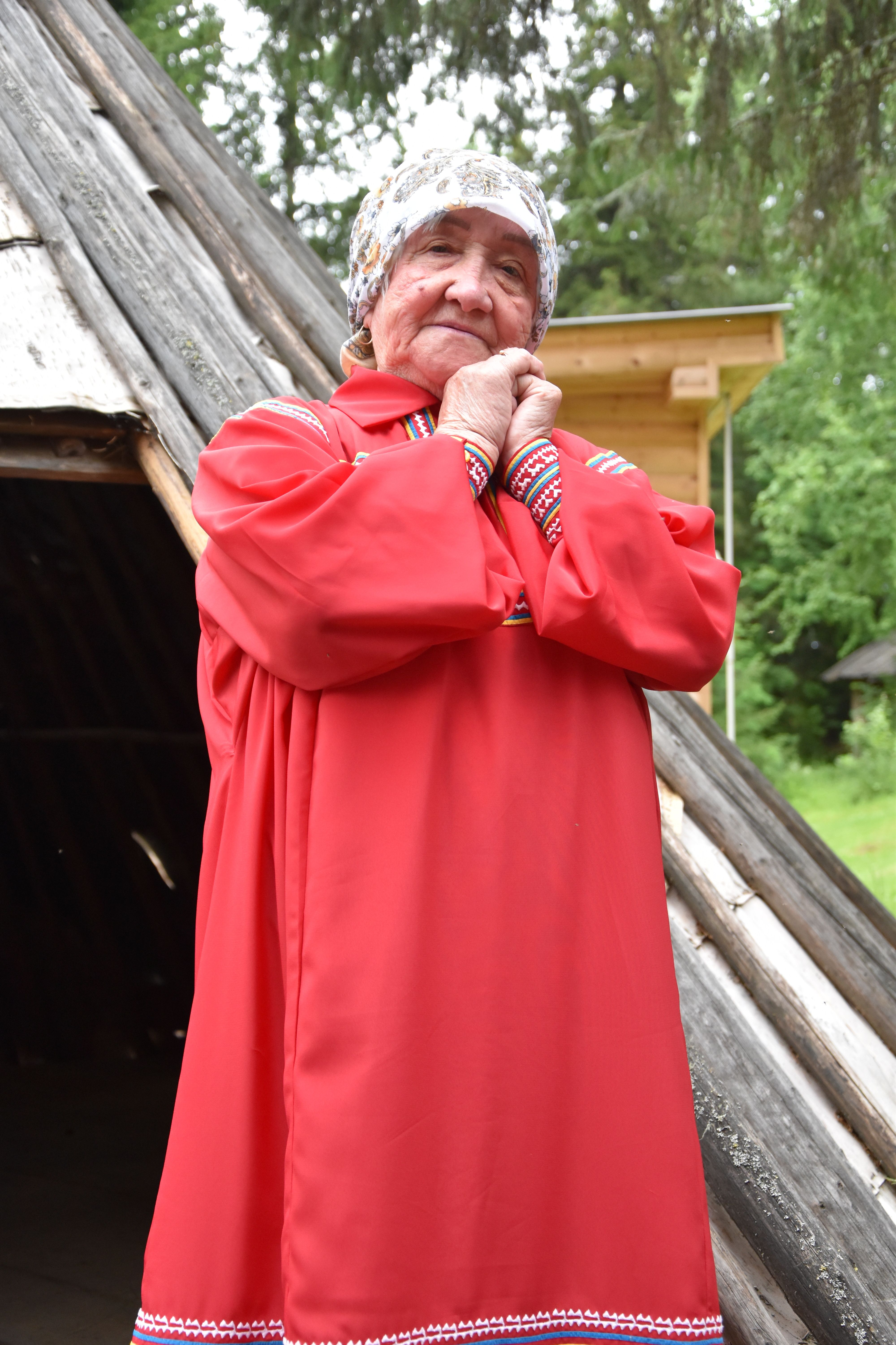 Portrait of an elderly woman in traditional red Russian dress outdoors, expressing warmth.