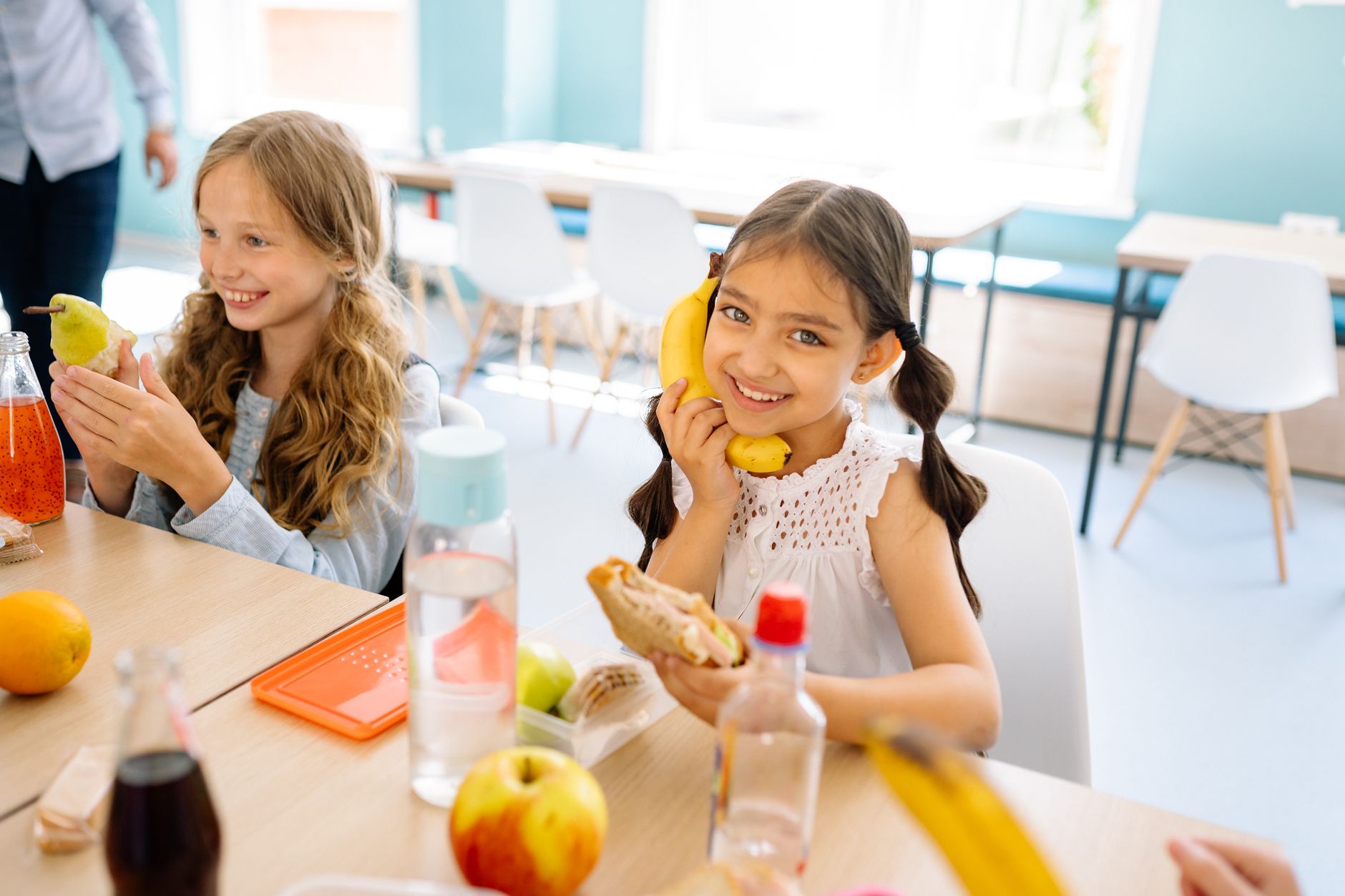 Two smiling girls enjoying snacks during break time in a school classroom.