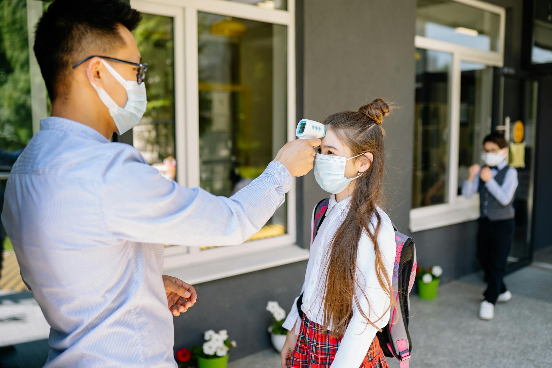 Masked teacher checks student's temperature with an infrared thermometer outside school.