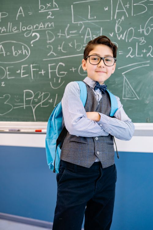 Free Young boy with glasses standing confidently in front of a math-filled chalkboard, embodying academic spirit. Stock Photo
