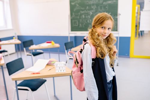 Free A young girl with a backpack stands confidently in a bright classroom setting. Stock Photo