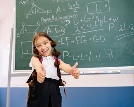 Smiling girl giving thumbs up in front of a chalkboard with math equations, wearing a white top.