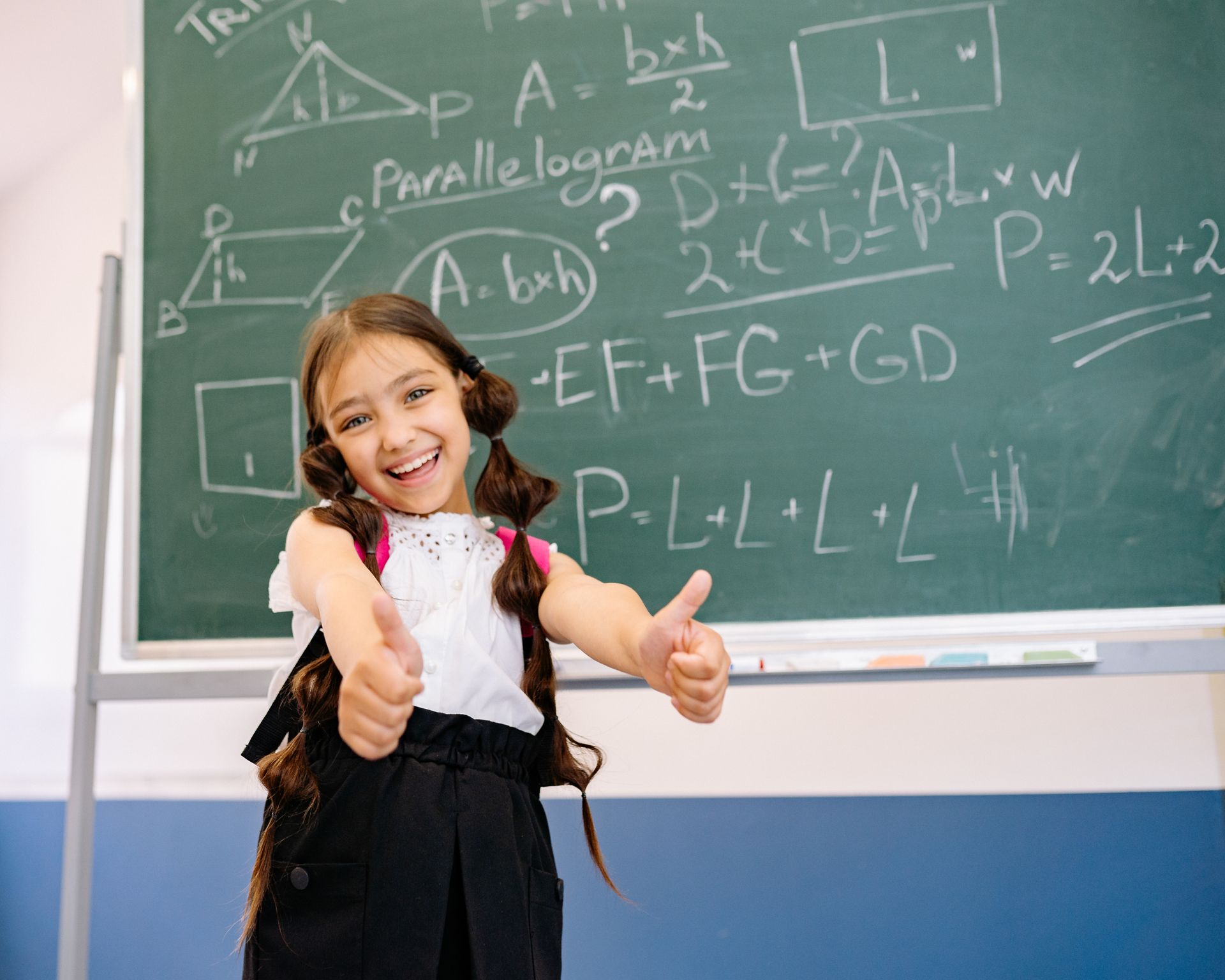 Smiling girl giving thumbs up in front of a chalkboard with math equations, wearing a white top.