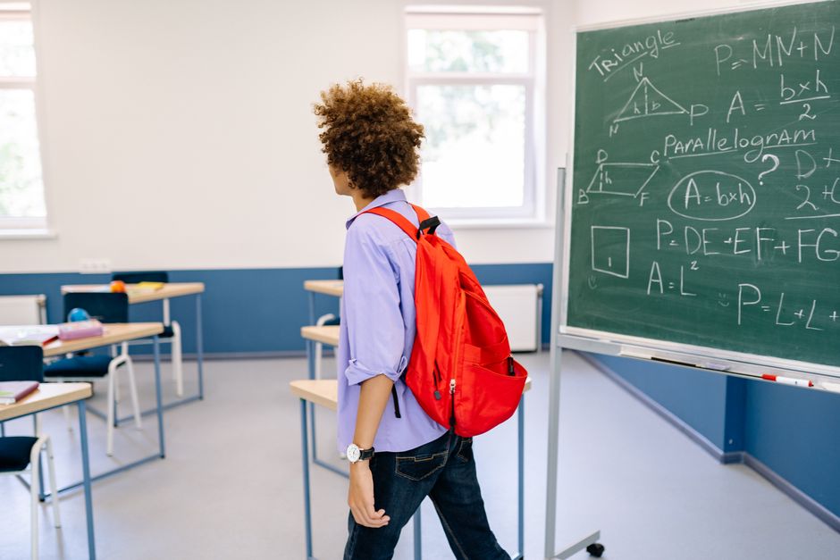 A student with a red backpack leaves a classroom with a chalkboard filled with math equations.