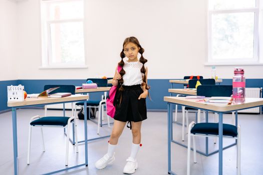 A young girl stands confidently in a bright, modern classroom with desks and educational materials.