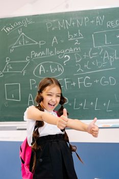 Cheerful girl in a classroom showing thumbs up with math formulas on the blackboard.