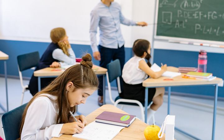 A teacher stands near a blackboard teaching students seated at desks in a classroom.