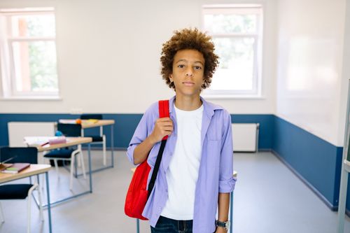 Free Young boy standing in a modern classroom, wearing a backpack, looking confidently at the camera. Stock Photo