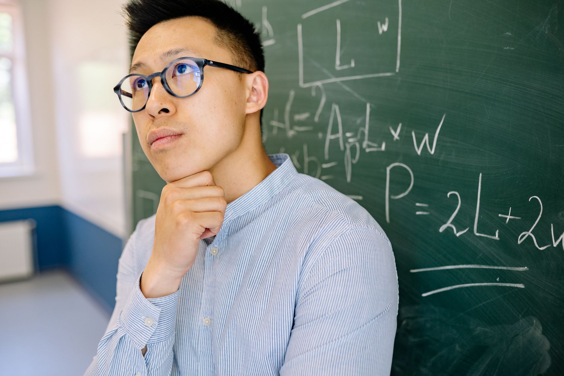 Educator in blue shirt contemplating mathematical equations on blackboard indoors.