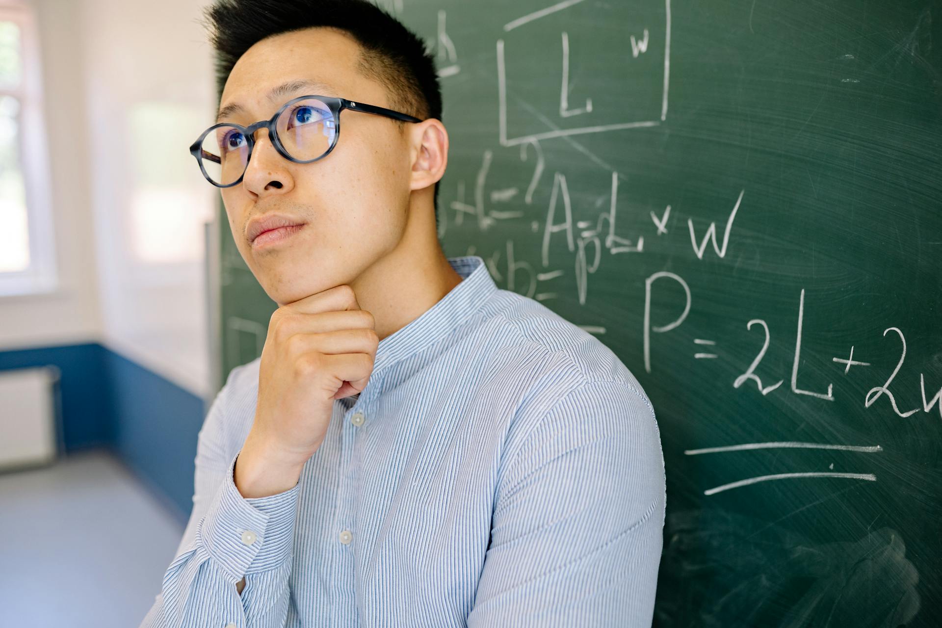 Educator in blue shirt contemplating mathematical equations on blackboard indoors.