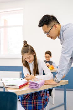 A teacher helps a student at her desk in a bright classroom environment.