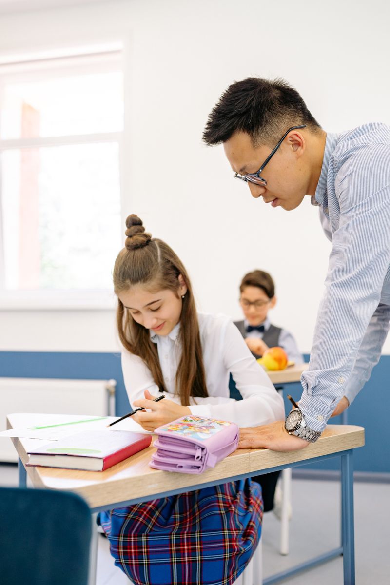 A teacher helps a student at her desk in a bright classroom environment.