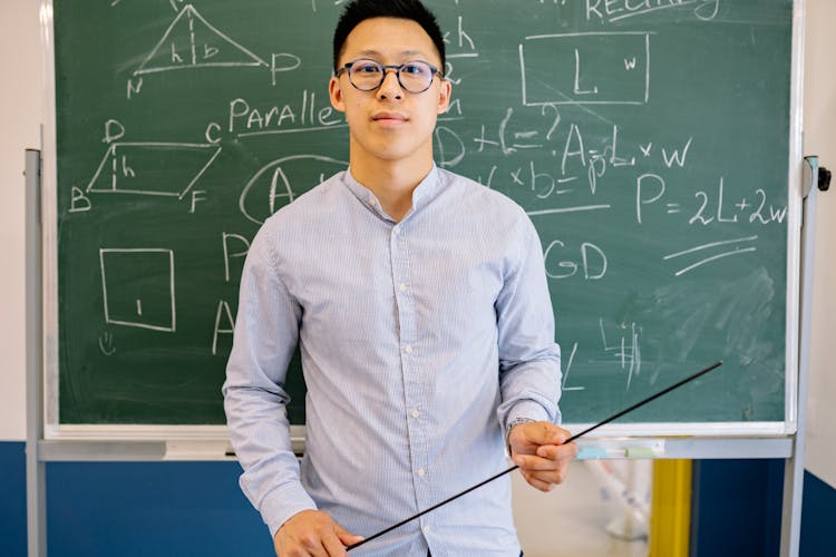 A Male Teacher Holding A Pointing Stick In Front Of A Blackboard