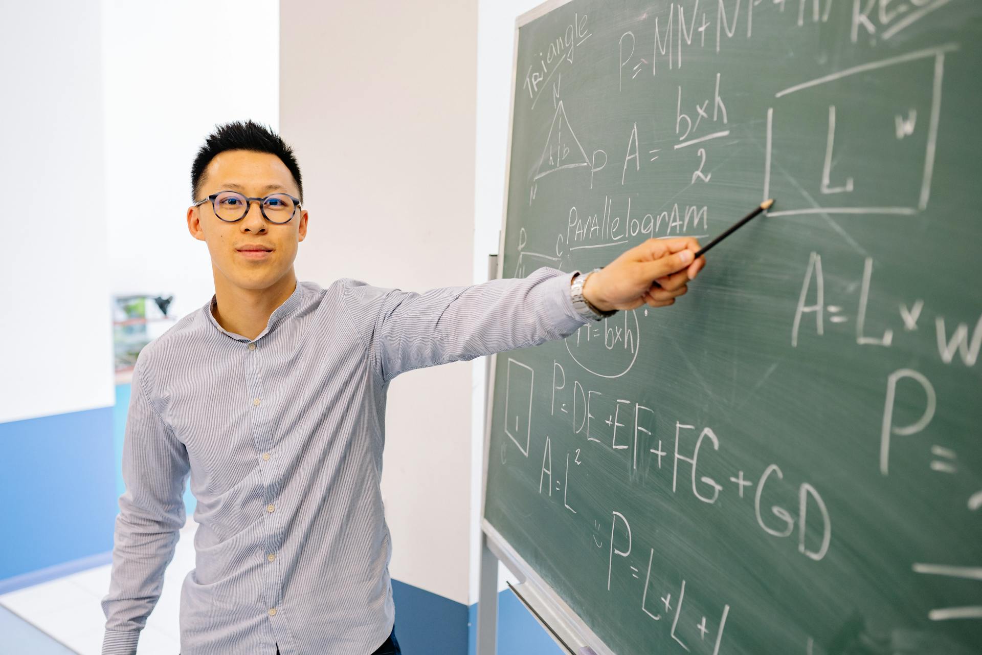 An Asian male teacher wearing eyeglasses points confidently at a blackboard filled with math equations in a classroom setting.