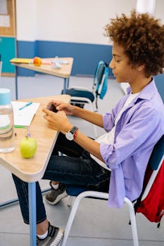 Teen boy engaged with smartphone during school break at desk.