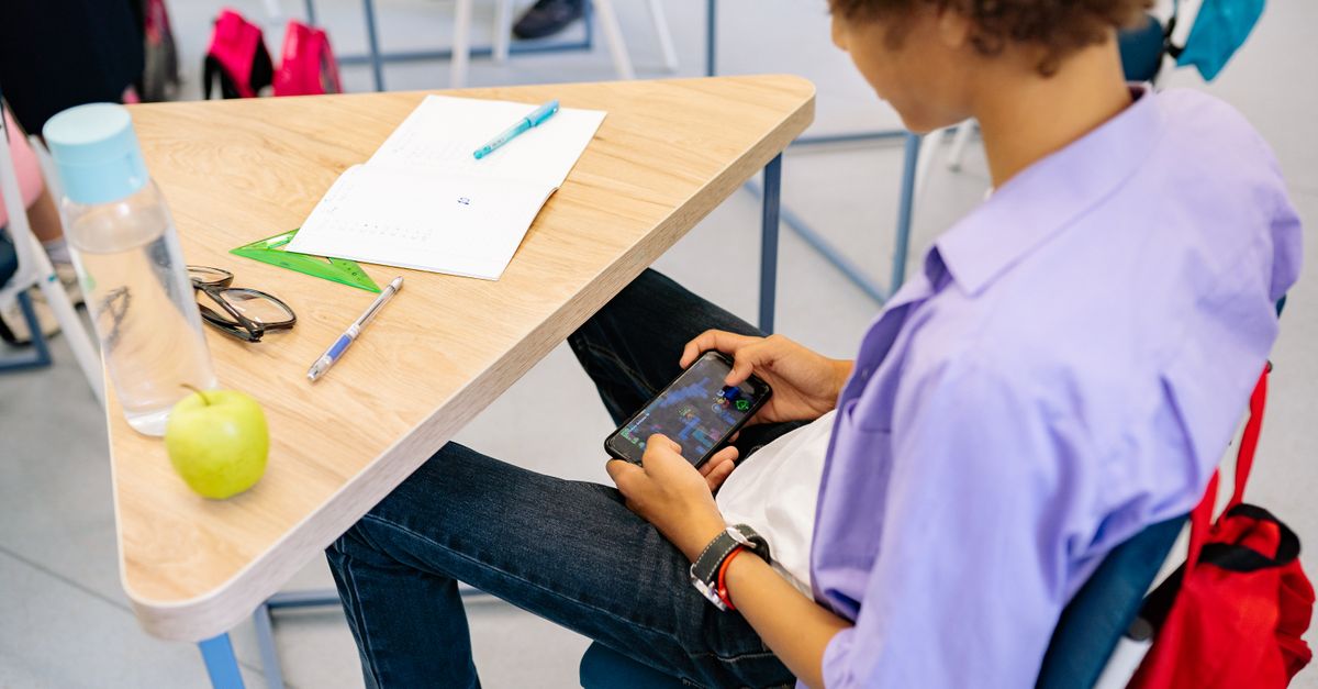 A child playing a mobile game at a school desk with school supplies nearby, captured indoors during class.